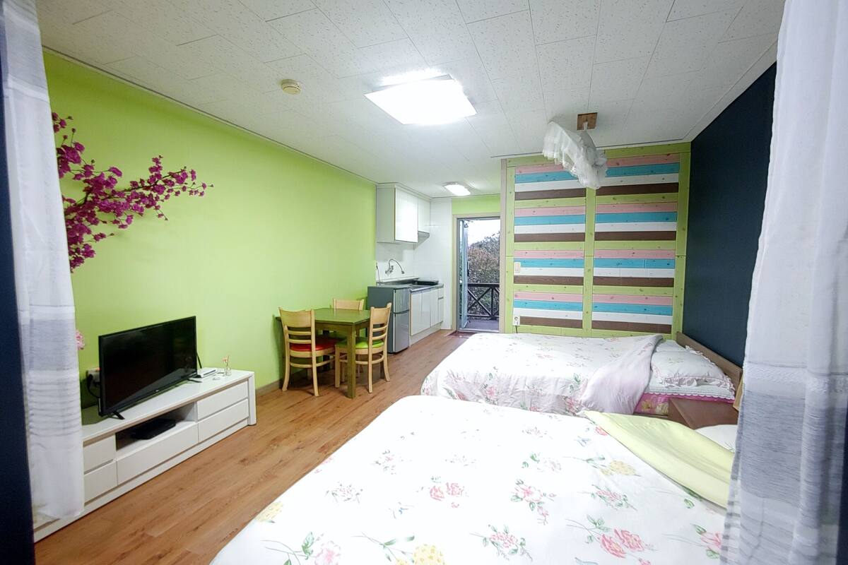 A room with two beds, a TV, a small table, and a kitchenette.