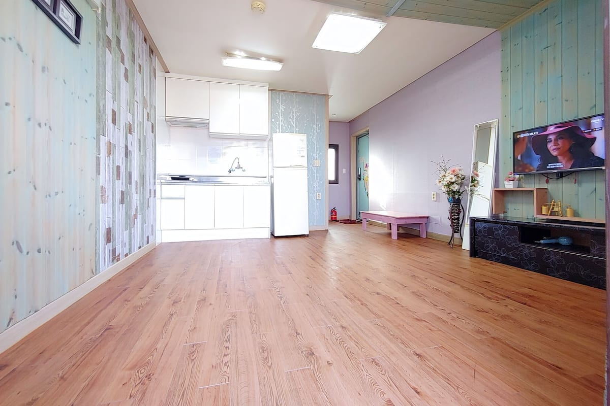 A spacious living area with a wooden floor, a kitchen with white cabinets, a bench, a TV, and a flower vase.