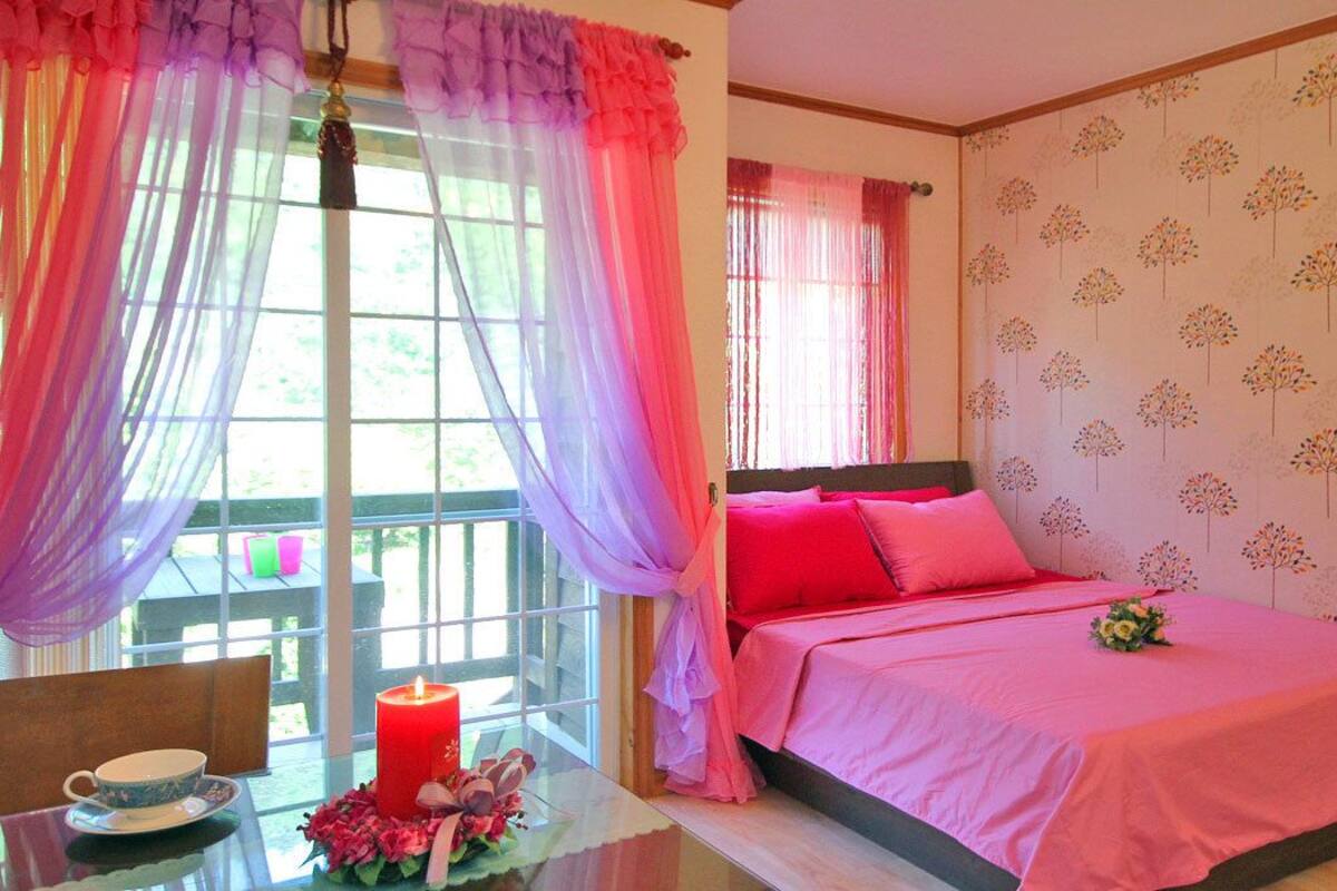 Un dormitorio con una cama, cortinas, una mesa con una taza y un platillo, y una vela roja.