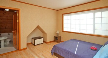 Un dormitorio con cama, ventana, una mesita y una puerta de madera que da a un baño.