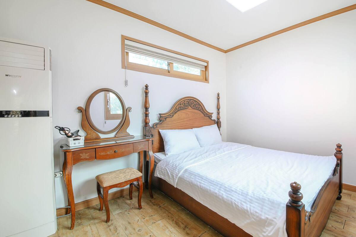 A bedroom with a wooden bed, a wooden dresser with a mirror, a bench, and a built-in heater.
