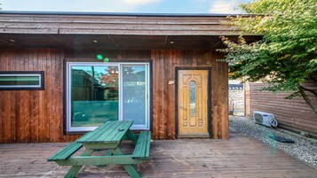 Un edificio de madera con un amplio ventanal y una puerta de madera, una mesa de picnic y un pequeño espacio al aire libre con un muro de piedra y un árbol.