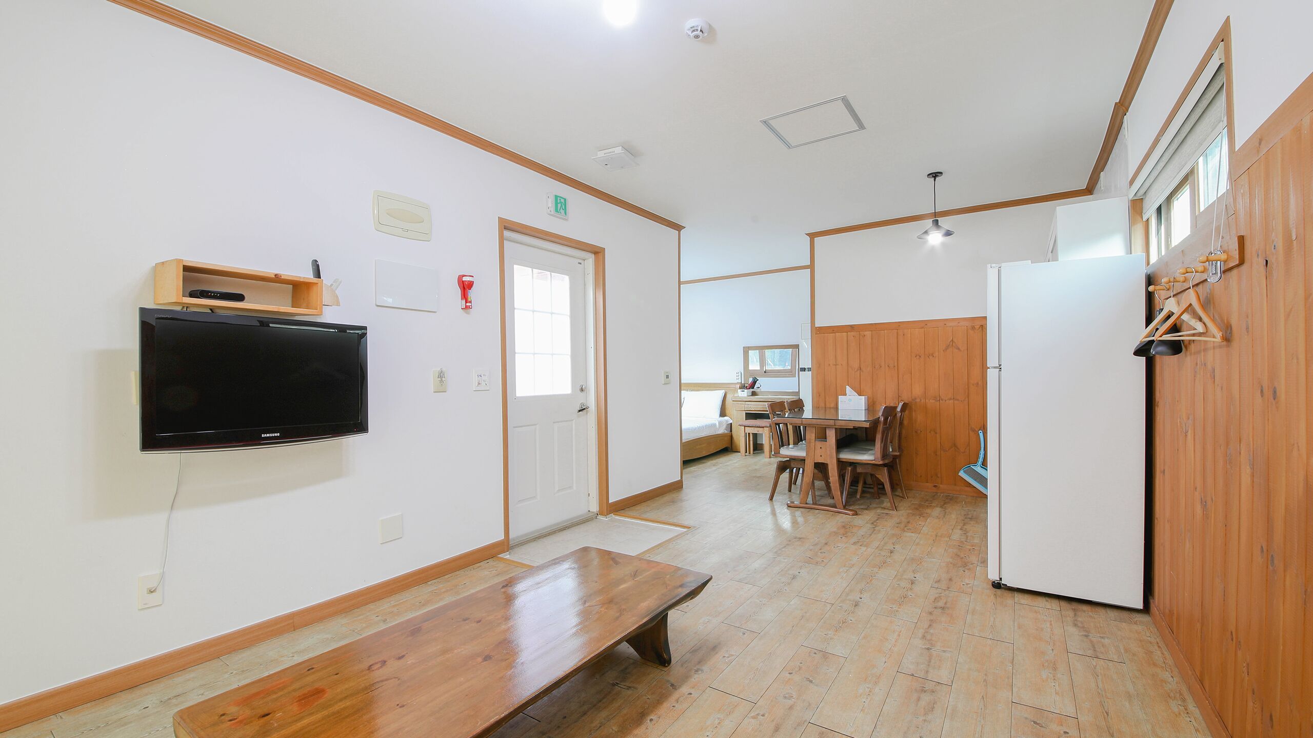 A room with a wooden bench, a TV mounted on the wall, a wooden dining table with chairs, and a refrigerator.