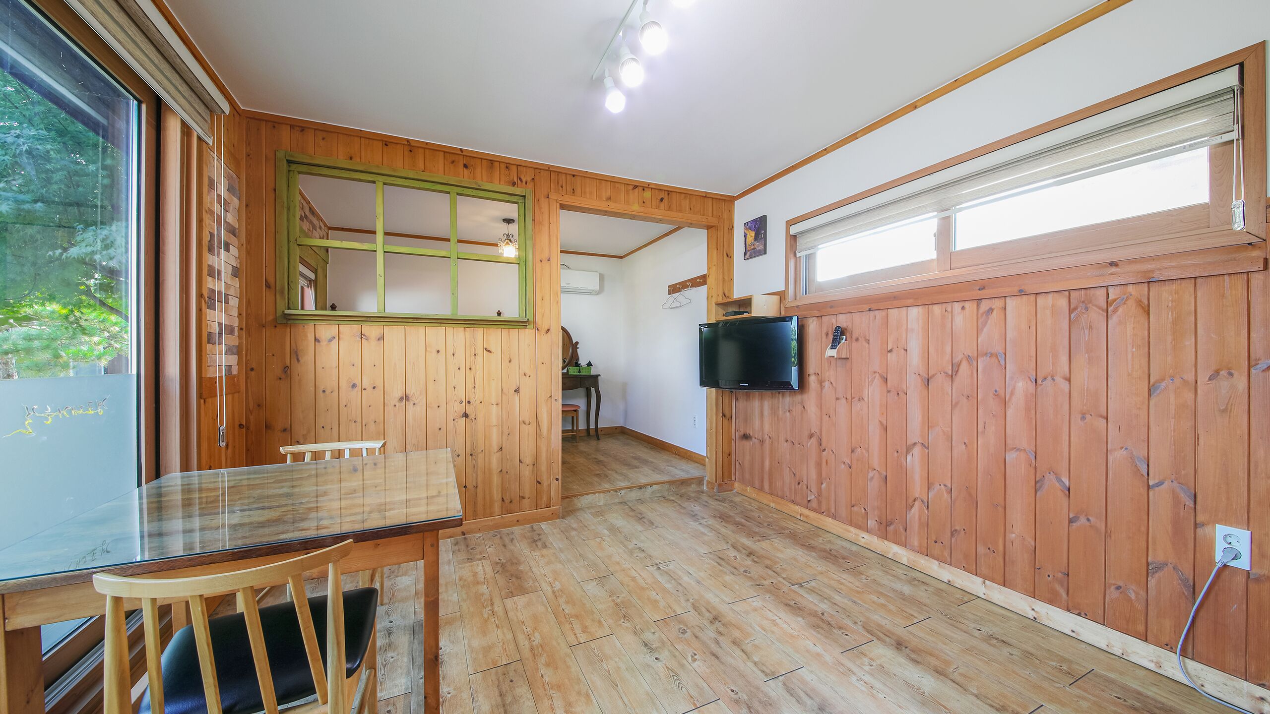 A wooden interior with a dining table, chairs, a TV mounted on the wall, and a window with blinds.