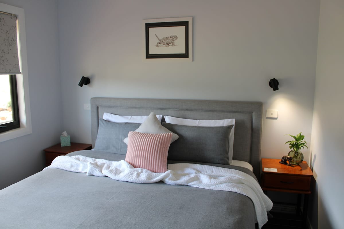 A neatly made bed with a grey headboard, a pink pillow, and a white blanket. There is a framed picture on the wall above the bed and a small plant on the nightstand beside the bed.