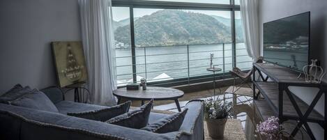 A modern living room with a large window overlooking a body of water and hills, a sofa, a small table, and a flat-screen TV.