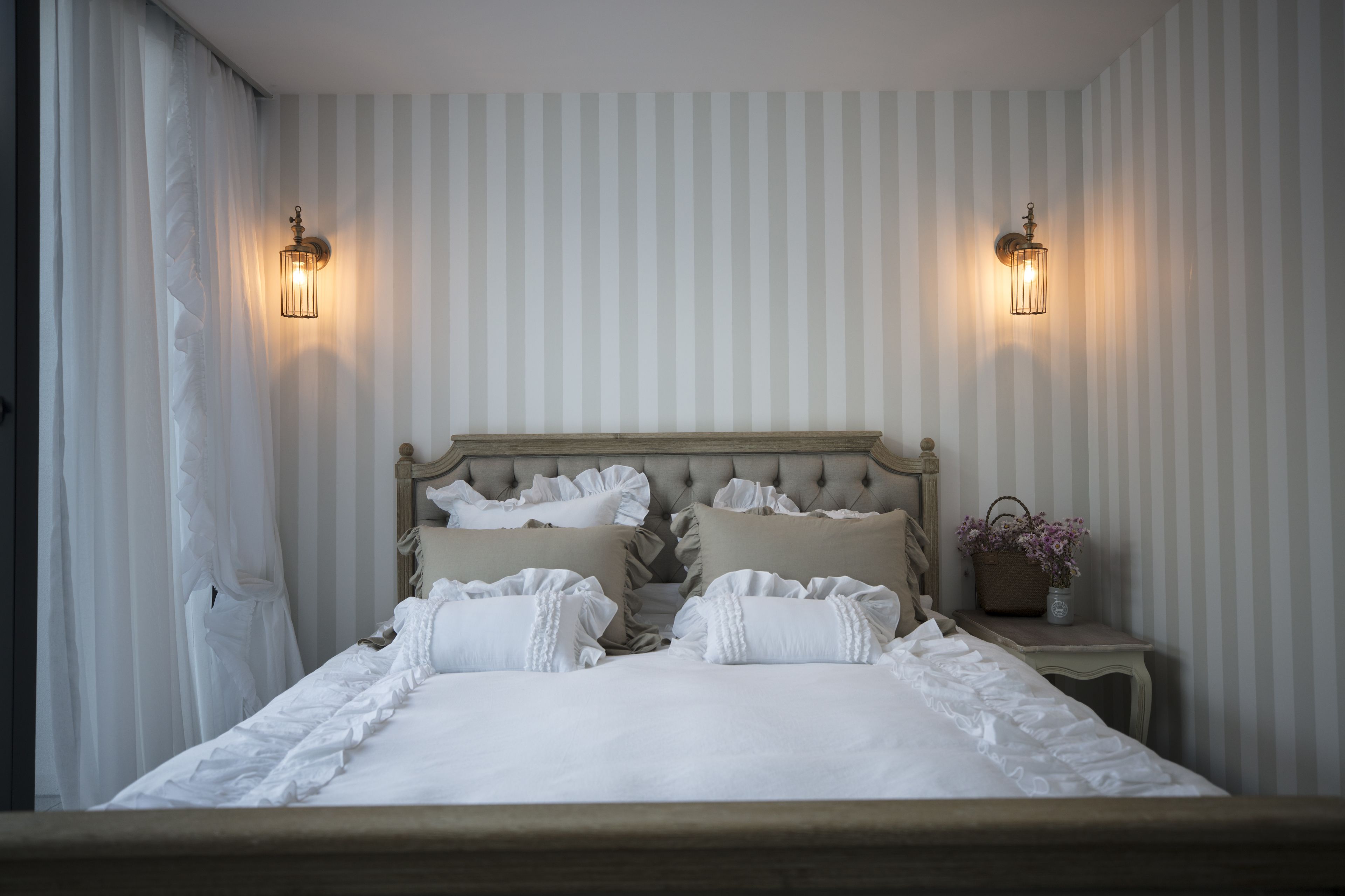 A neatly made bed with white linens and ruffled pillows, flanked by two wall sconces.