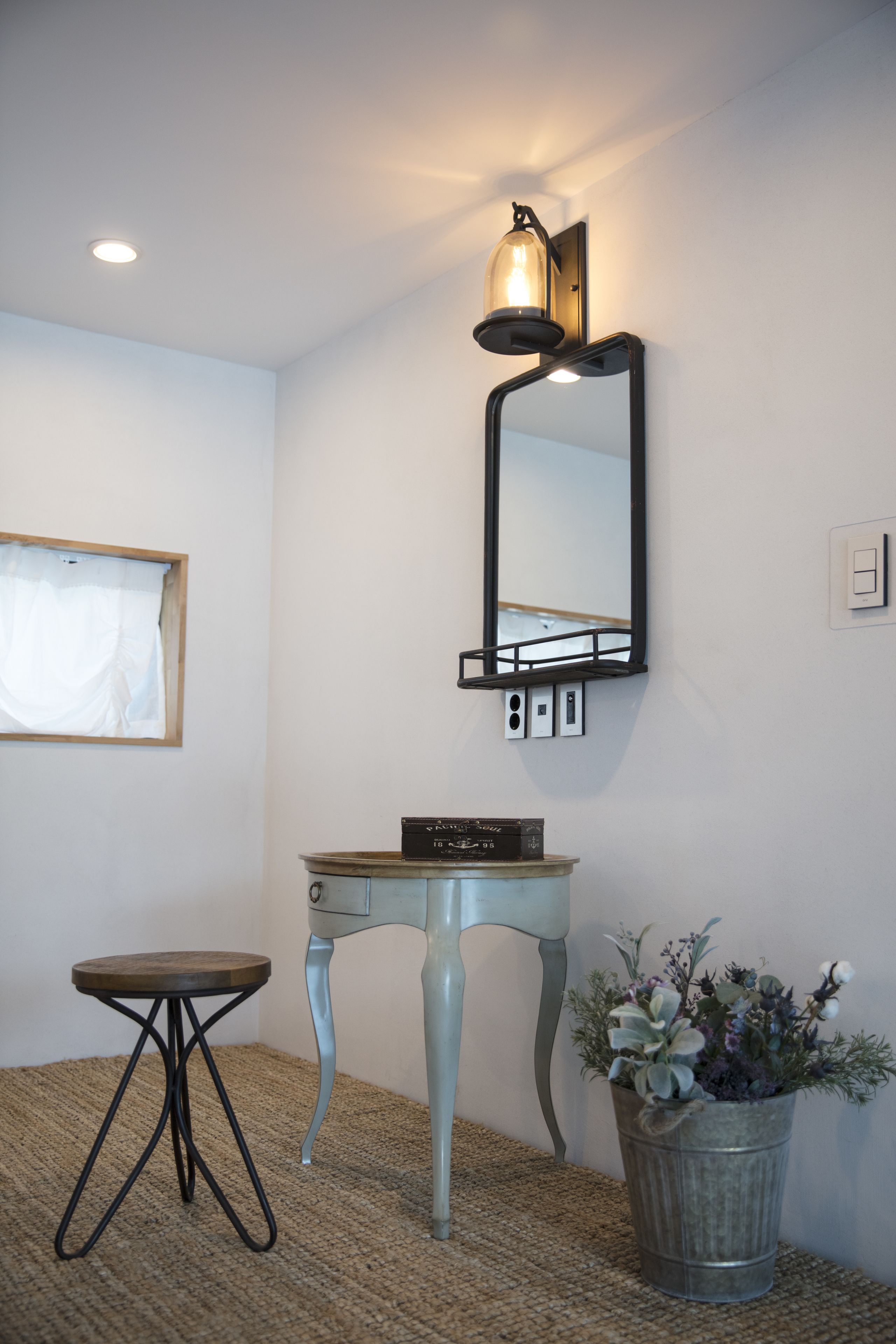 A bathroom with a vanity, a mirror, a wall-mounted light, a small table, and a potted plant.
