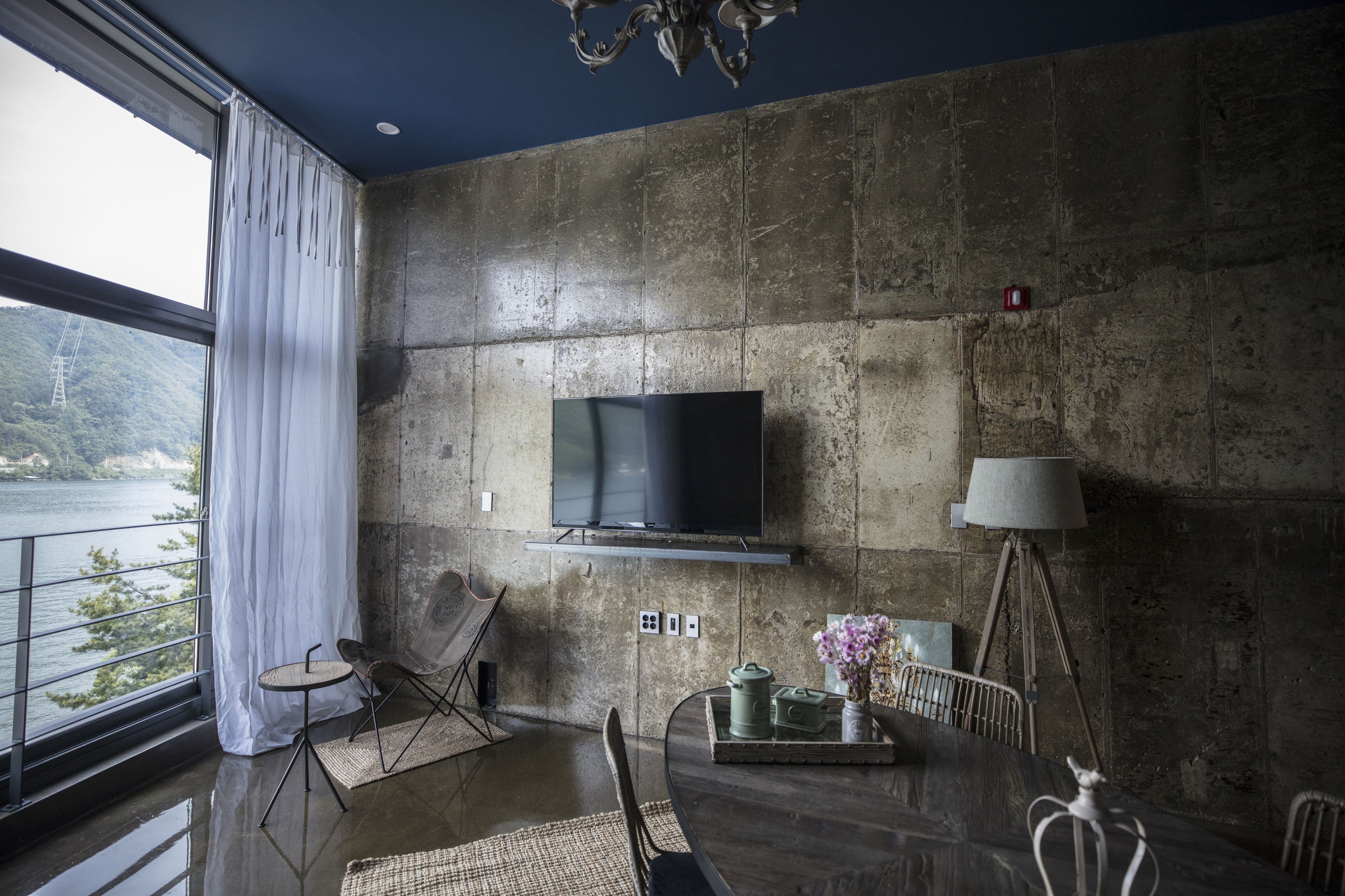 Een moderne woonkamer met een groot raam, een flatscreen-televisie bevestigd aan een gestructureerde muur, een eettafel met stoelen en een lamp.
