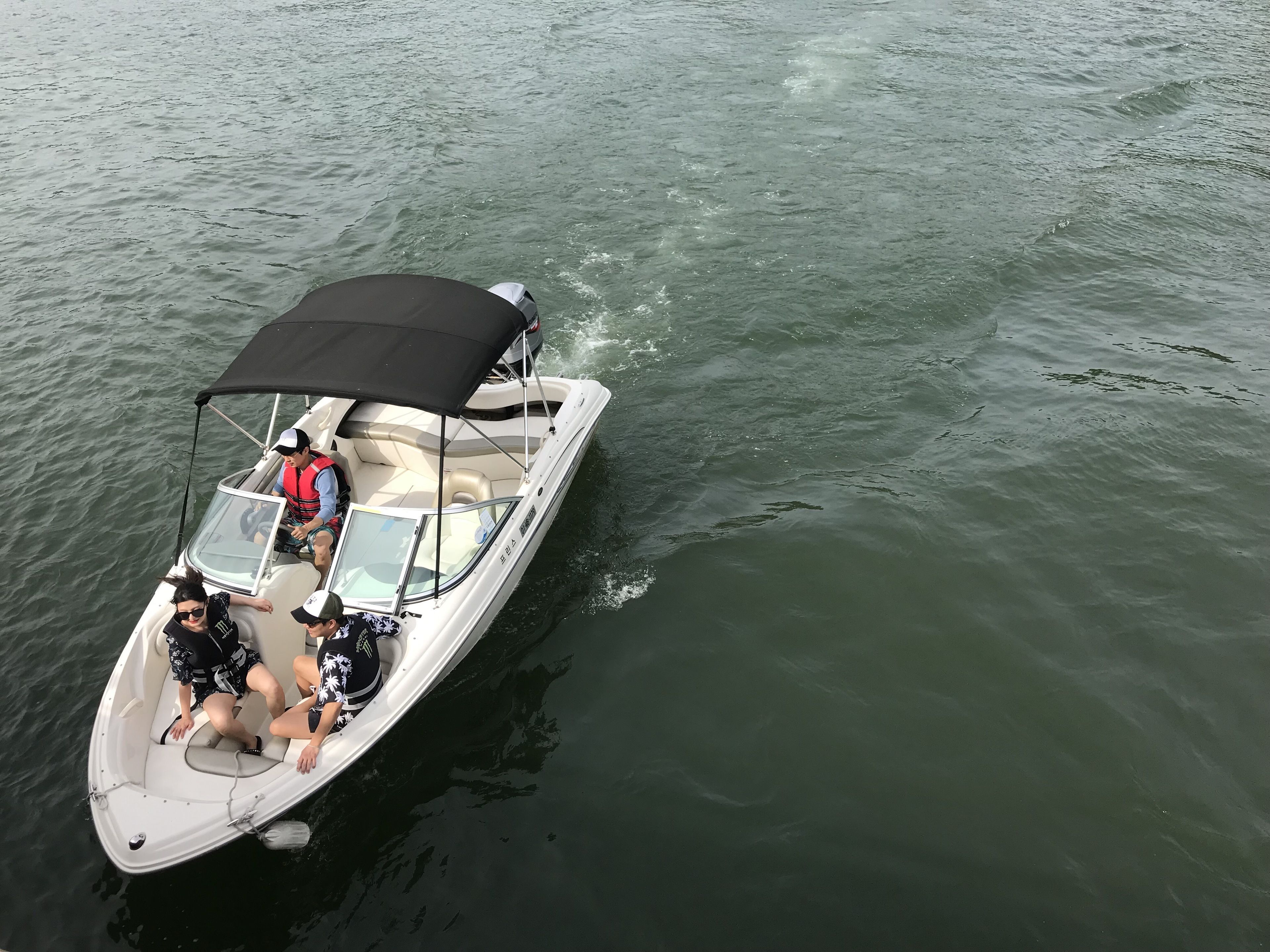 A small motorboat with a black canopy, two people on board, one wearing a life jacket, and a third person seated at the back.