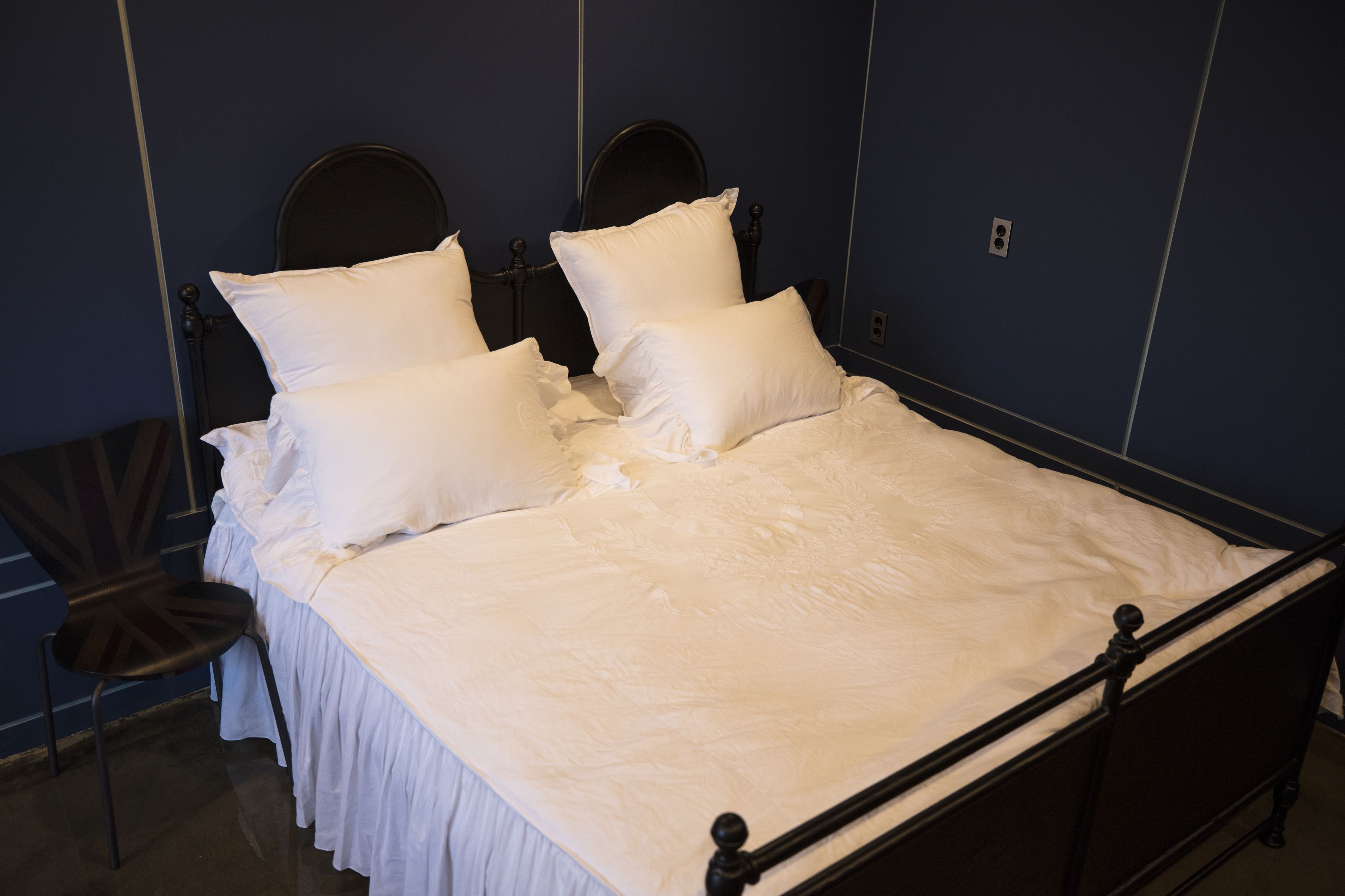 Un lit bien fait, avec du linge de lit blanc et des oreillers, un cadre en métal foncé et une chaise dotée d’un coussin foncé.