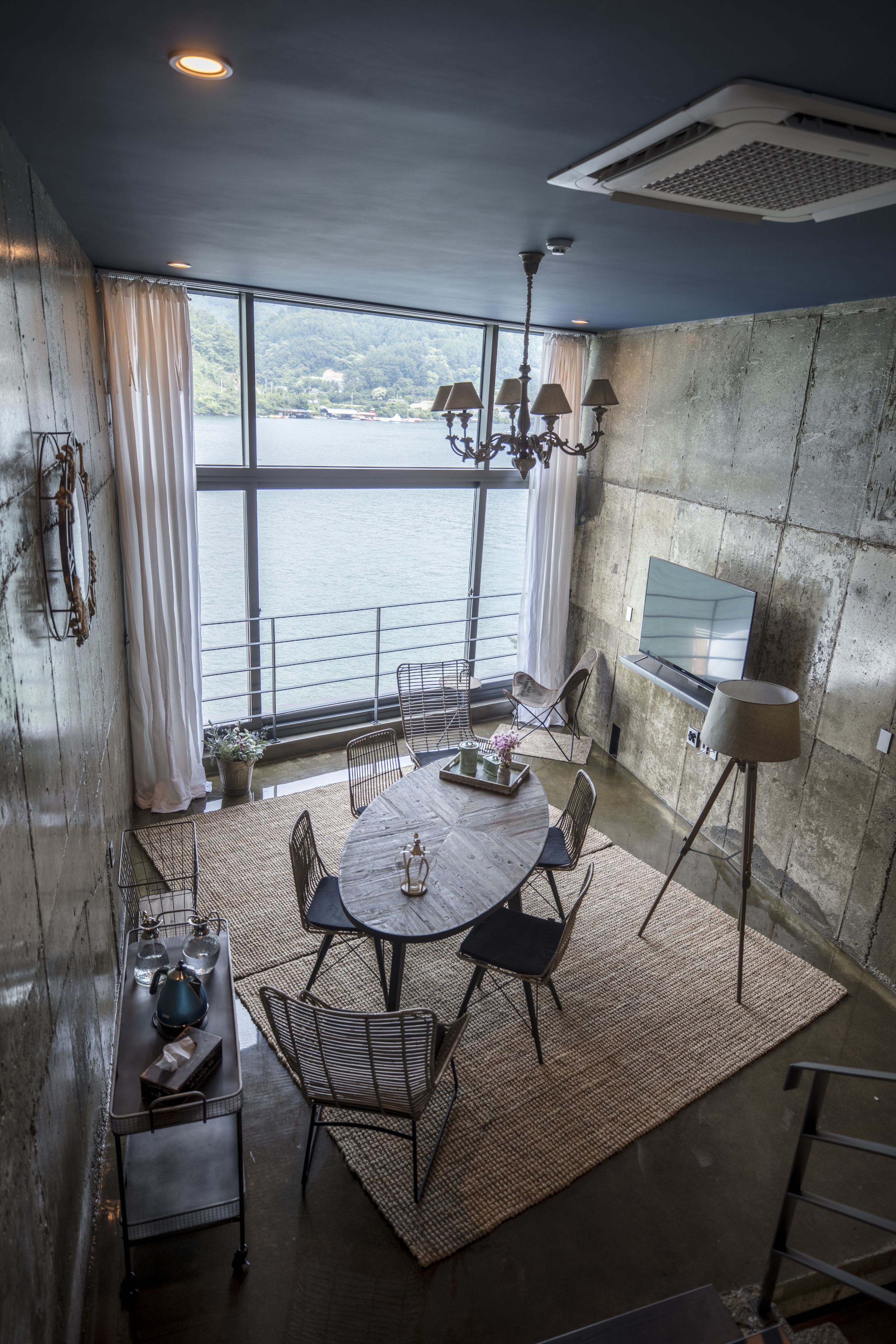 A dining area with a round table, chairs, and a chandelier. There is a TV mounted on the wall and a window with a view of the water.
