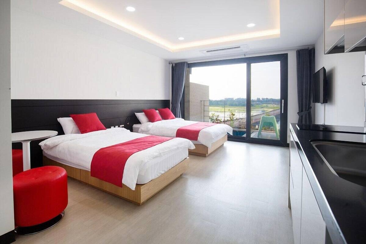 Habitación de hotel con dos camas, una mesa de comedor, zona de cocina y un amplio ventanal corredero que da a un balcón.