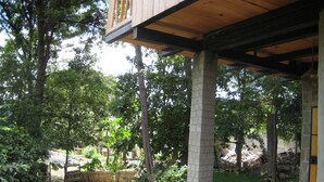 A covered outdoor area with a wooden deck, stone pillars, and a view of a garden with trees and a fence. - Tzikin Jaay (San Pedro La Laguna)