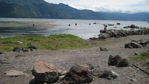 A rocky shoreline with a person standing near the water's edge, a distant mountain range, and a clear sky. - Tzikin Jaay (San Pedro La Laguna)