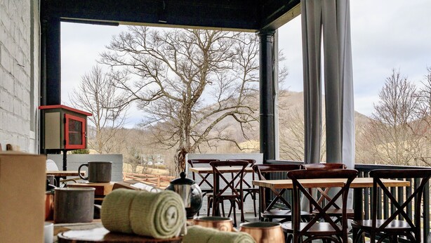 A rustic dining area with wooden tables and chairs, a large window offering a view of bare trees and mountains, and various kitchen utensils and cookware on the tables. - The Inn At Warm Springs (Warm Springs)