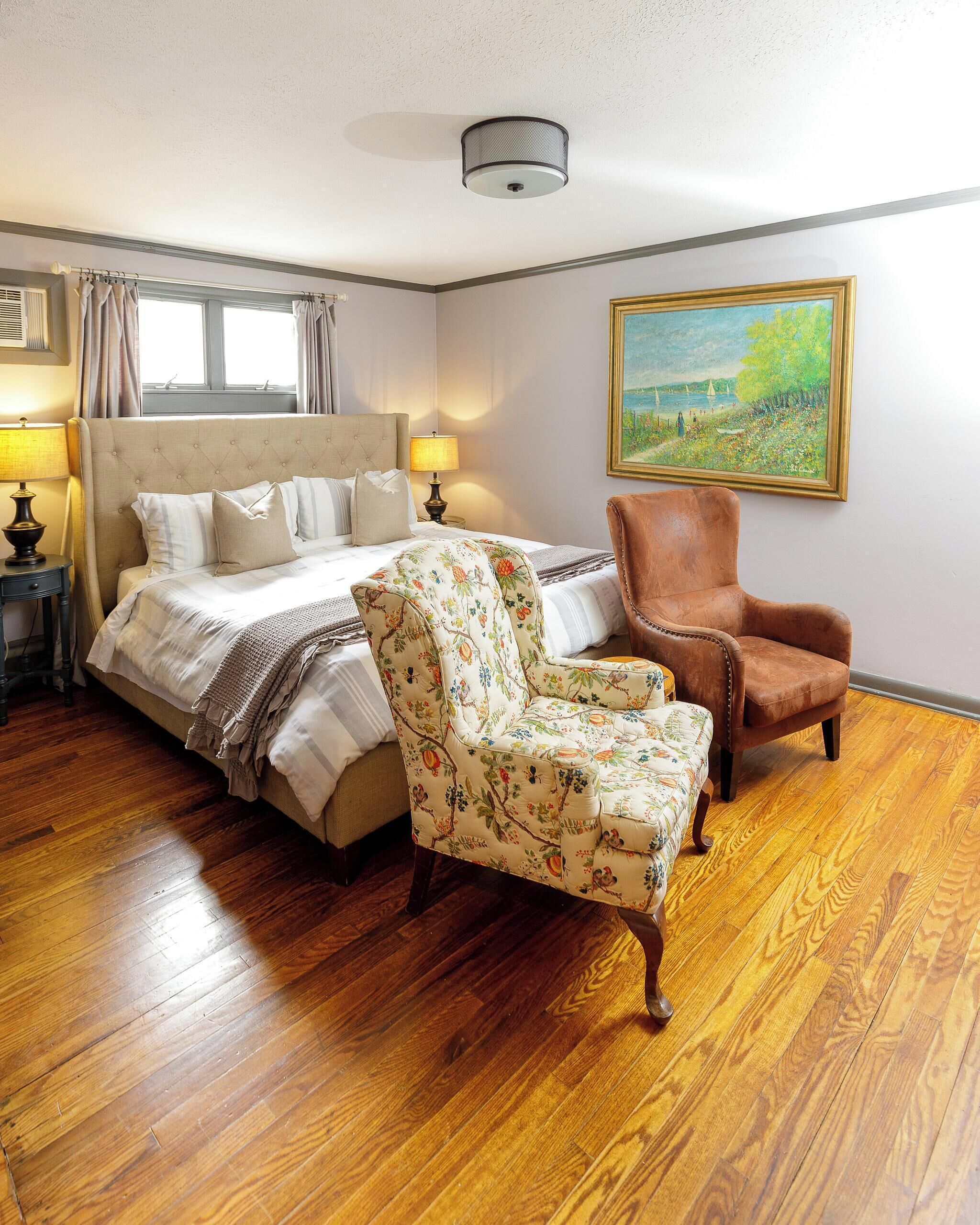 A bedroom with a large bed, a floral armchair, a brown leather chair, wooden flooring, a painting on the wall, and a ceiling light fixture.