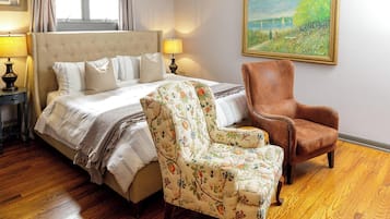 A bedroom with a large bed, a floral armchair, a brown leather chair, wooden flooring, a painting on the wall, and a ceiling light fixture.