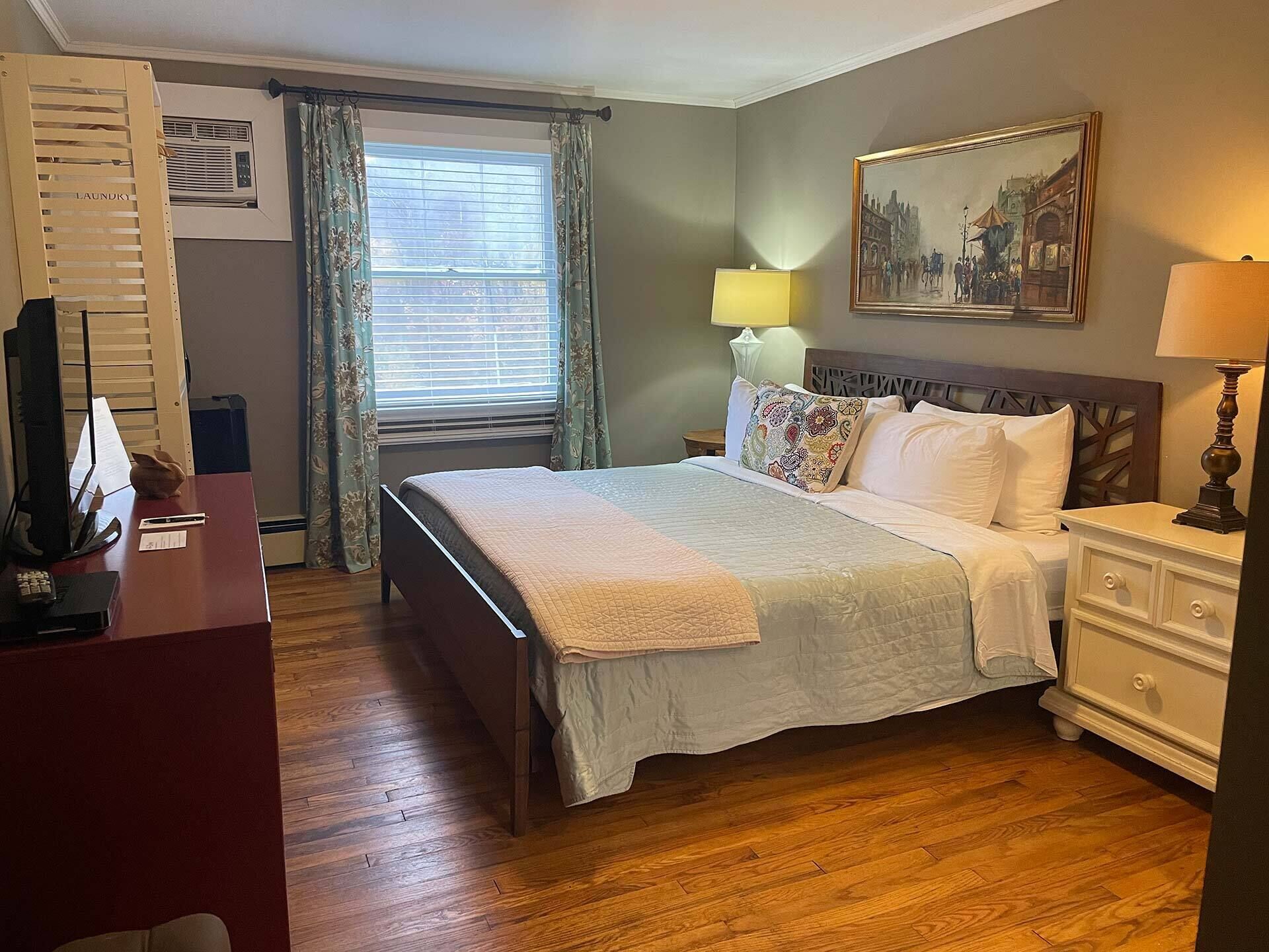 A bedroom with a bed, bedside tables, a desk, a lamp, a painting, and a window with curtains.