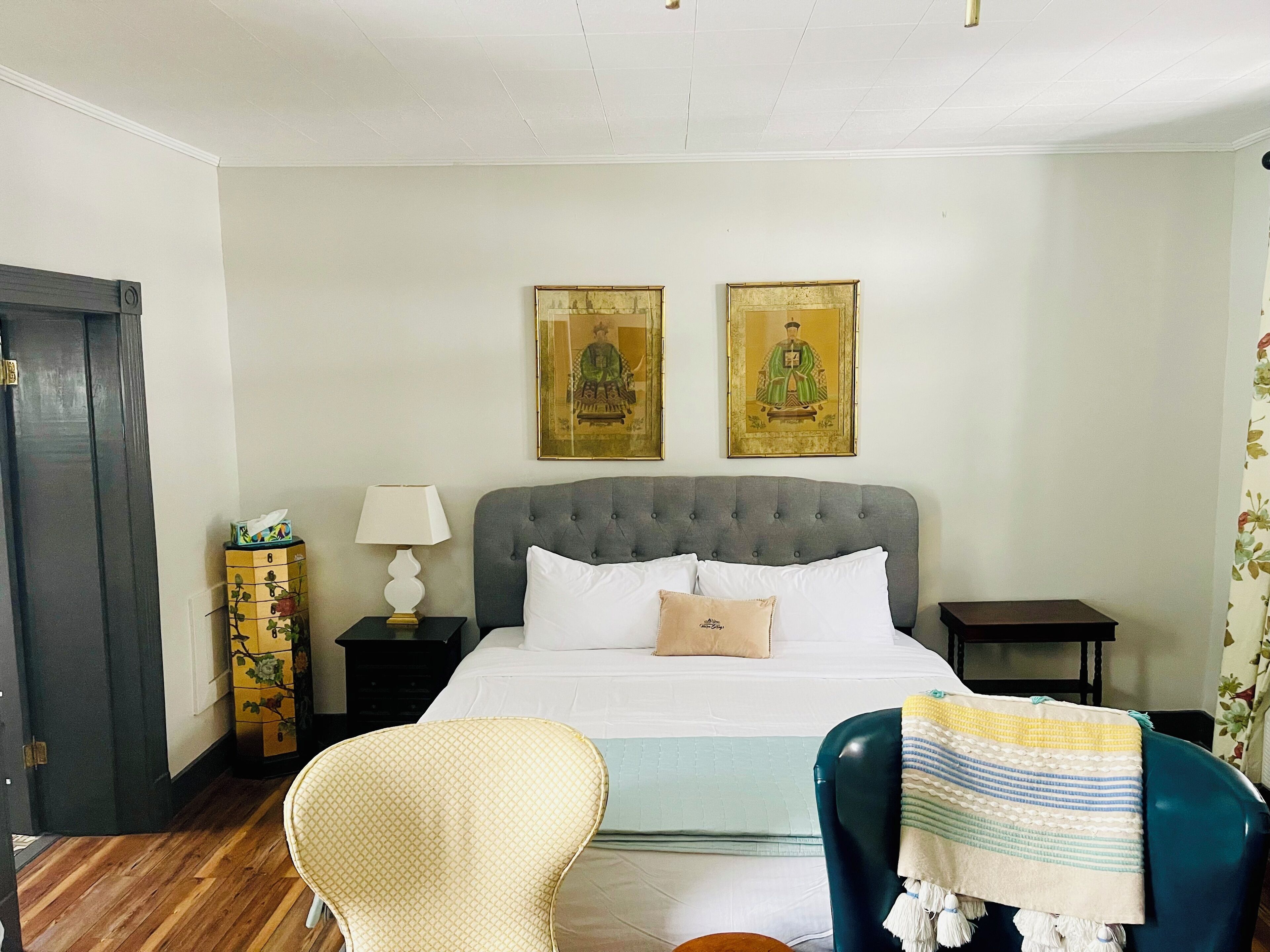 A neatly arranged bedroom with a bed, two framed artworks on the wall, a bedside lamp, a chair, and a small table.