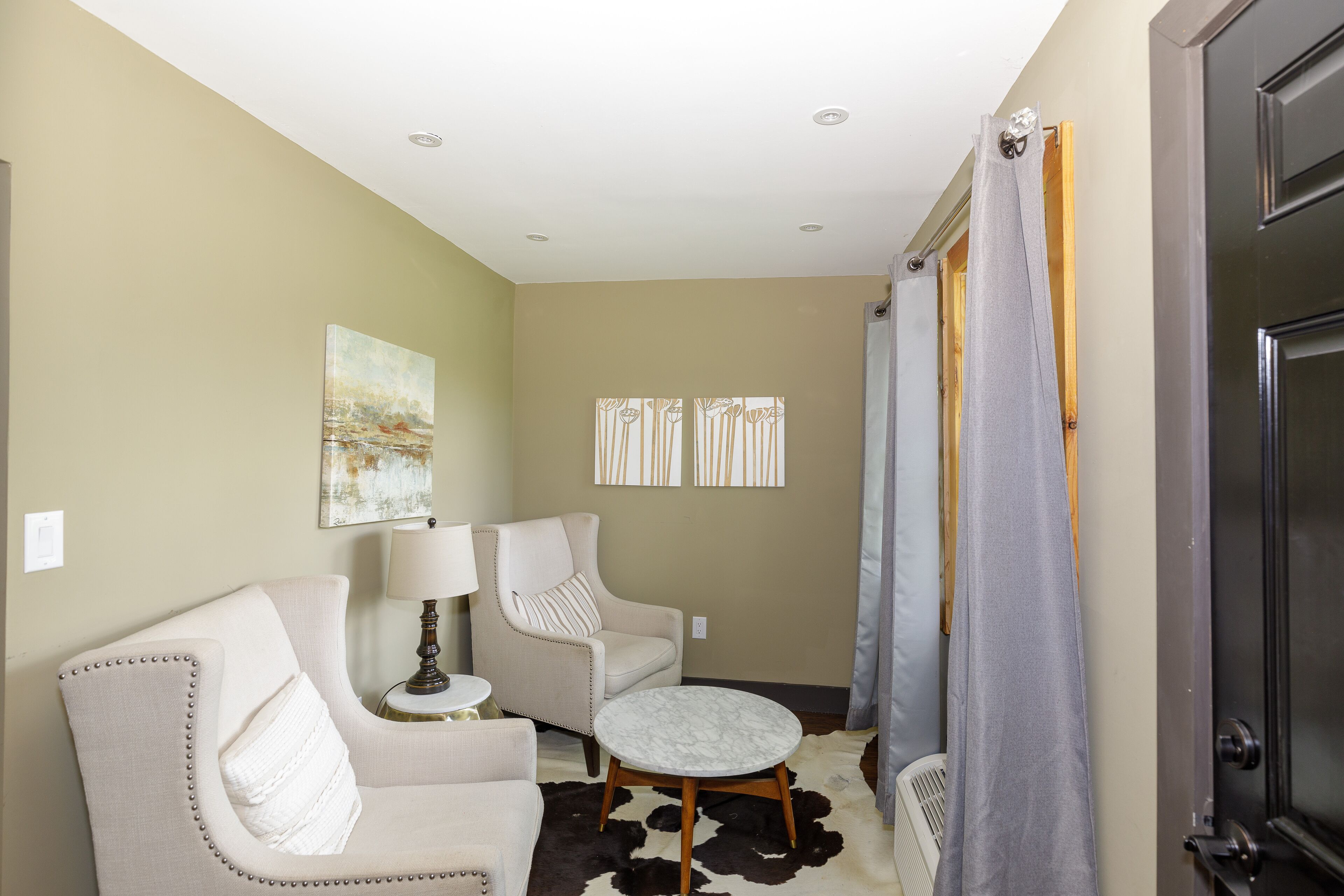 A living room with a beige sofa, a round coffee table, and a chair. There is a painting on the wall and a window with striped curtains.