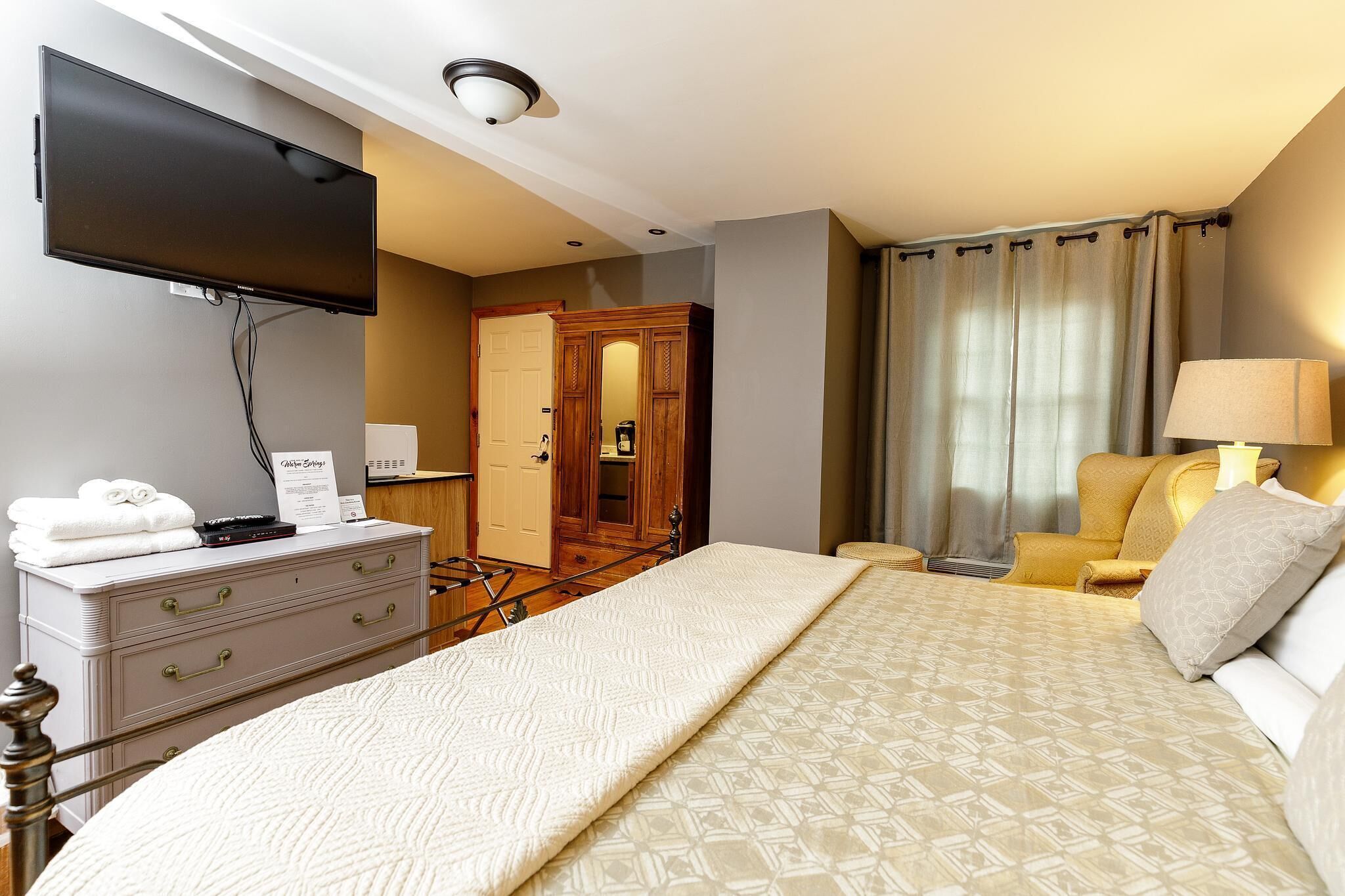Habitación de hotel con cama, cómoda, televisor de pantalla plana en la pared, una silla y una lámpara.