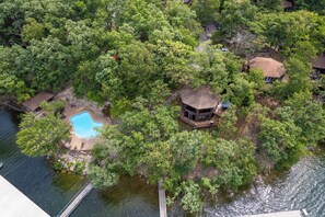 Vista aérea de una propiedad aislada con piscina, terraza de madera y construcción de techo de paja rodeada de abundante vegetación.