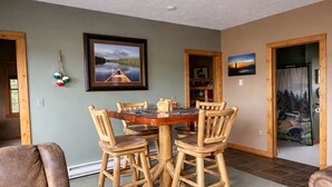 A dining area with a wooden table and chairs, a framed picture on the wall, and a door with a forest scene curtain. - The River's Edge Hot Tub Wi-Fi Riverfront Pet Nature Relax Ski Stargaze Relax!  (Indian River)