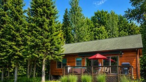 A wooden cabin with a deck, surrounded by a well-maintained lawn and tall trees. - The River's Edge Hot Tub Wi-Fi Riverfront Pet Nature Relax Ski Stargaze Relax!  (Indian River)