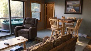 A living room with a sofa, a coffee table, a TV, a fireplace, a dining table with chairs, and a hot tub outside. - The River's Edge Hot Tub Wi-Fi Riverfront Pet Nature Relax Ski Stargaze Relax!  (Indian River)