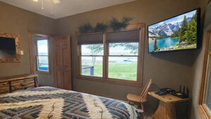 A bedroom with a large bed, a wooden dresser, a chair, a small table, a TV, and a window with a view of a lake and mountains. - The River's Edge Hot Tub Wi-Fi Riverfront Pet Nature Relax Ski Stargaze Relax!  (Indian River)