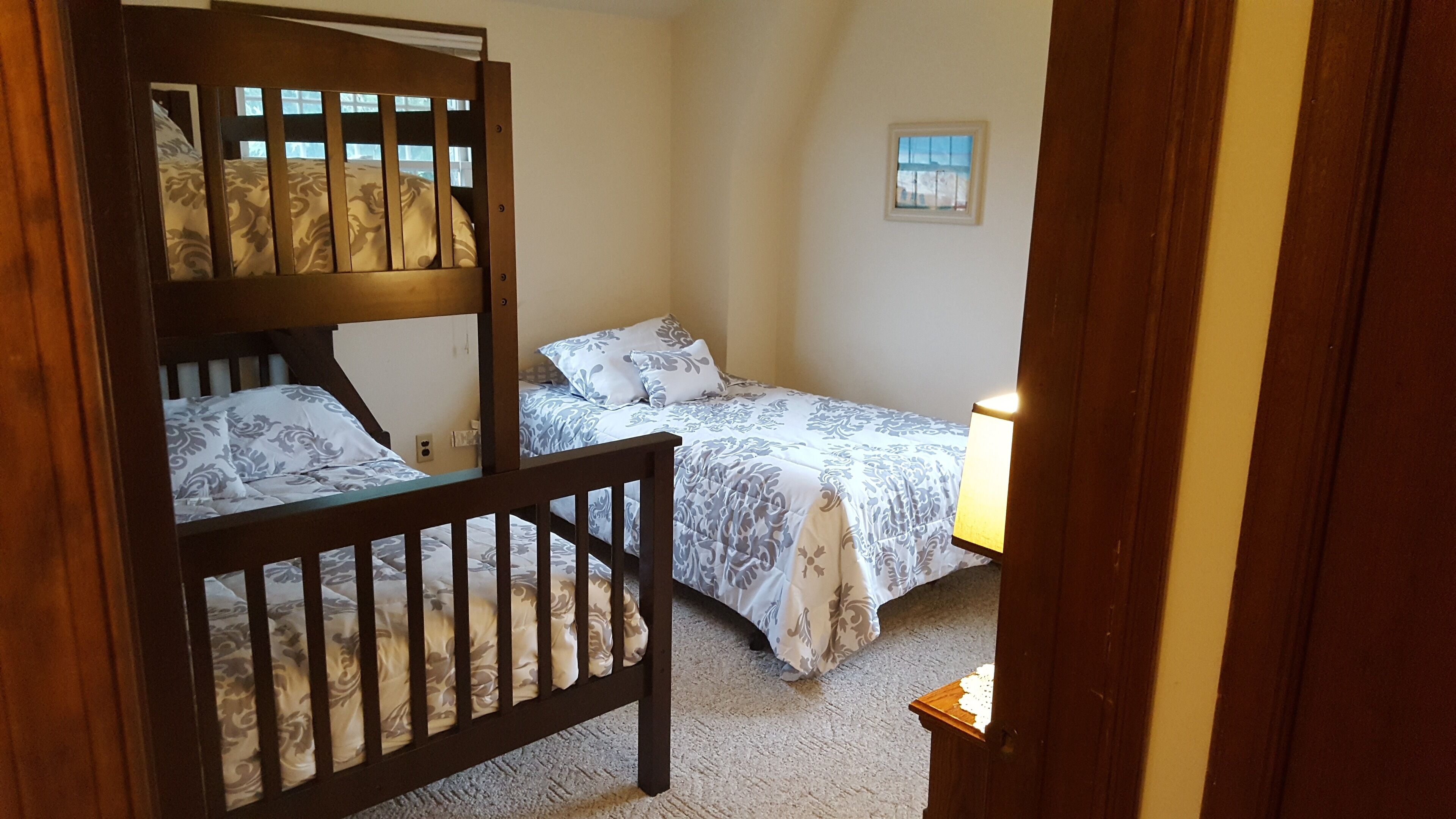 A room with two bunk beds and a single bed, each with patterned bedding. A small table with a lamp is visible.