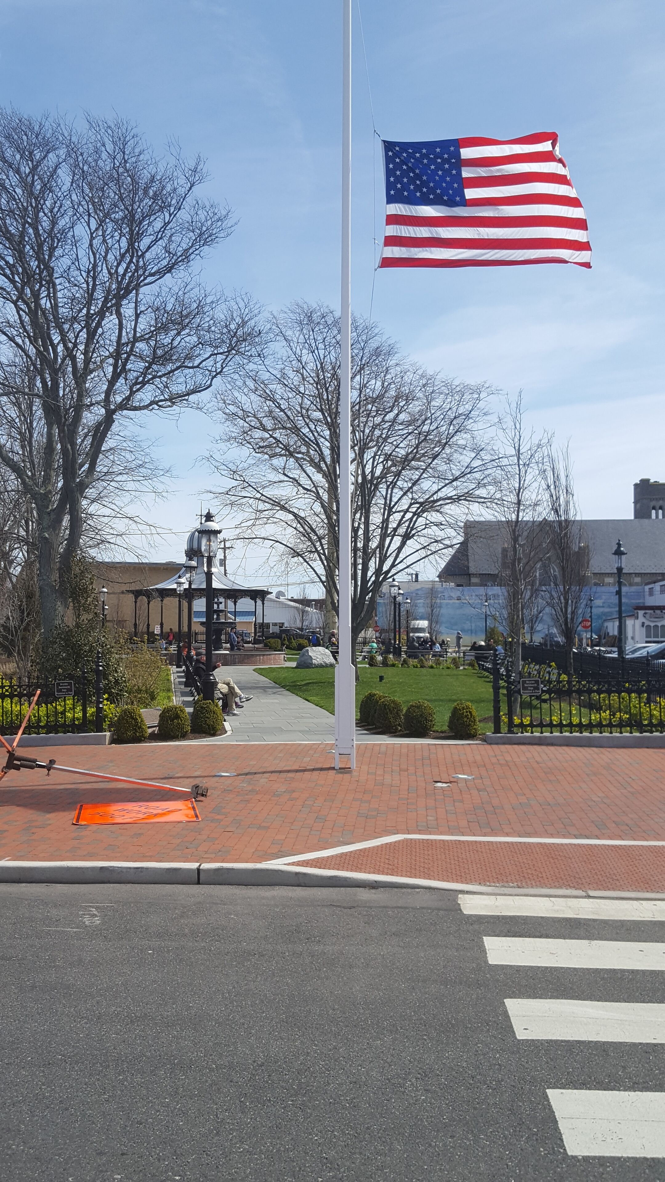 An American flag on a flagpole in a public square with a paved walkway, benches, and a lamp post.