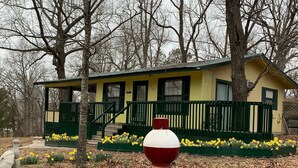A yellow house with green trim and a porch, surrounded by trees and daffodils. - Beaver Lake Rent Cabin  Lg Covered Deck Room~Firepit~Quiet~Private~7Mi Rogers AR (Rogers)