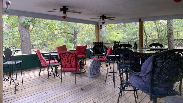 A covered outdoor seating area with wooden flooring, red and black chairs, a table, and ceiling fans. - Beaver Lake Rent Cabin  Lg Covered Deck Room~Firepit~Quiet~Private~7Mi Rogers AR (Rogers)