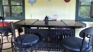 A covered outdoor dining area with a glass table and black chairs, a red blender, and a 'Certified Wildlife Habitat' sign on the wall. - Beaver Lake Rent Cabin  Lg Covered Deck Room~Firepit~Quiet~Private~7Mi Rogers AR (Rogers)
