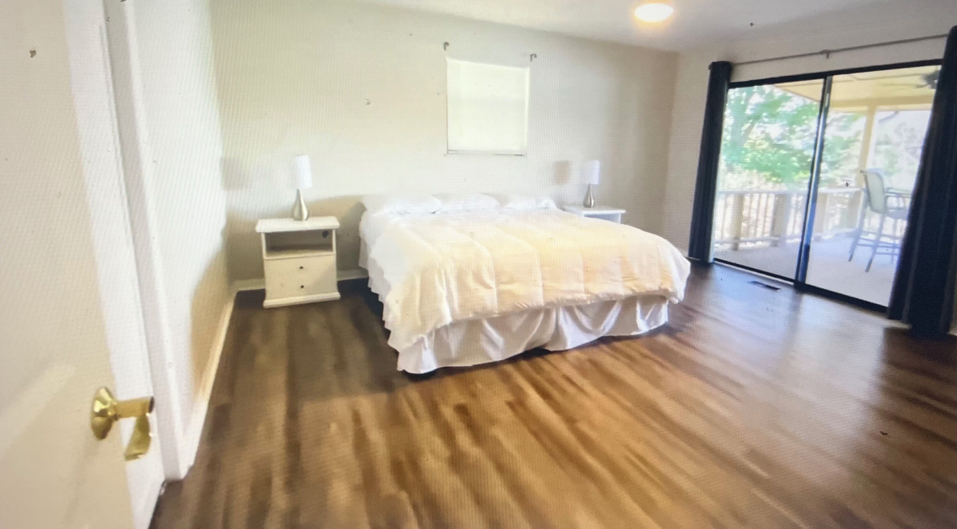Un dormitorio con suelo de madera, cama con ropa de cama blanca, mesita de noche, lámpara y vista a través de un ventanal.