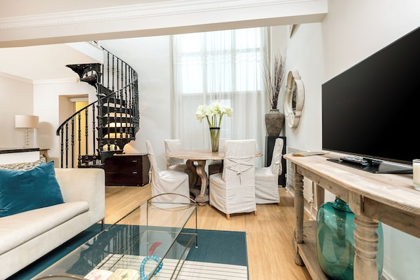 A modern living room with a staircase, a dining area with white chairs, a TV, and a large window with sheer curtains.