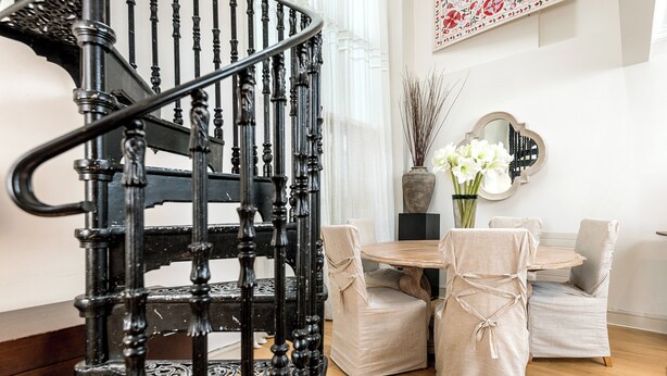 Una escalera con barandillas negras decorativas, una mesa redonda de madera con dos sillas y un jarrón con flores blancas.