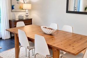 Un comedor con una mesa de madera, sillas blancas, un cuenco blanco, una lámpara y un cuadro en la pared.