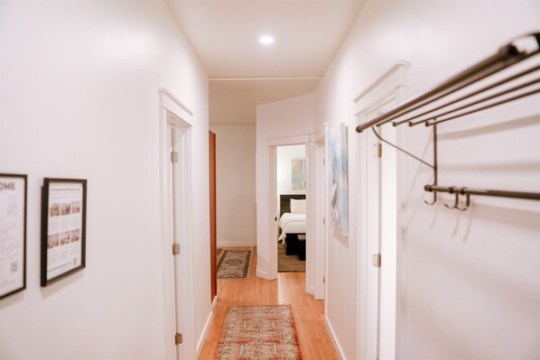 A hallway with a wooden floor, white walls, and a hanging towel rack. There are framed pictures on the wall and a rug on the floor.