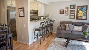 Una cocina moderna con refrigerador, microondas y taburetes de bar. Hay un comedor con mesa y sillas, y una sala de estar con sofá y una mesa de centro.