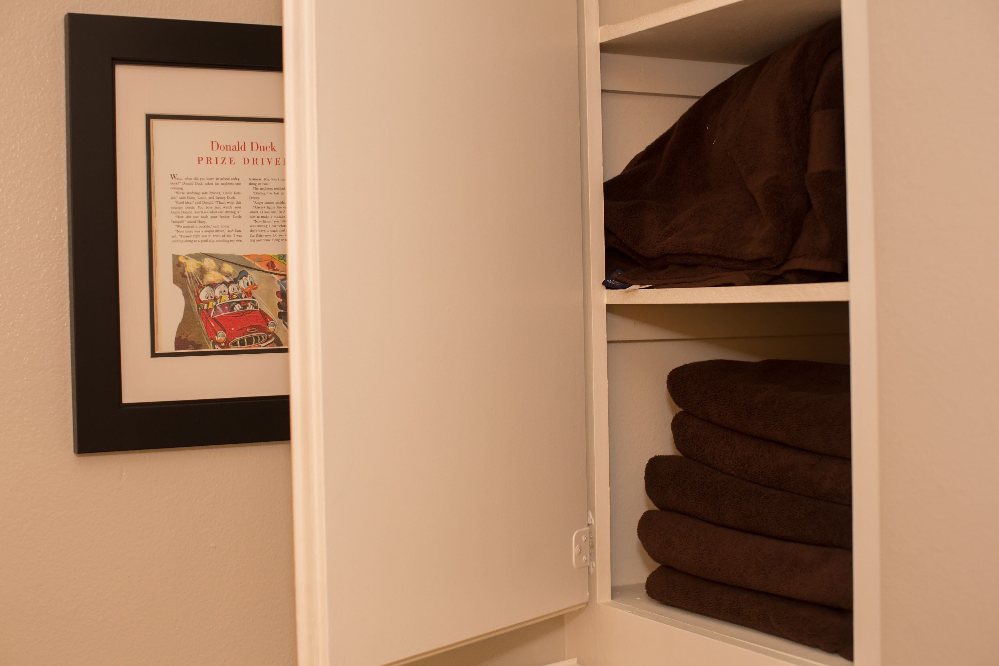 A framed picture of Donald Duck Prize Driver on a wall, a closet with folded towels, and a shelf with a brown item.