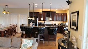 A modern kitchen with dark wood cabinets, a central island with bar stools, pendant lighting, and a dining area with a sofa and chairs.