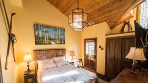 A bedroom with a wooden ceiling, a bed with white linens, a painting of a forest, and a wooden door.