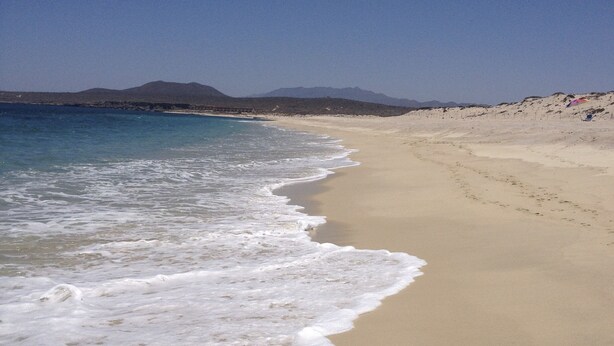 Una platja amb cel blau, mar tranquil·la i muntanyes a la distància.