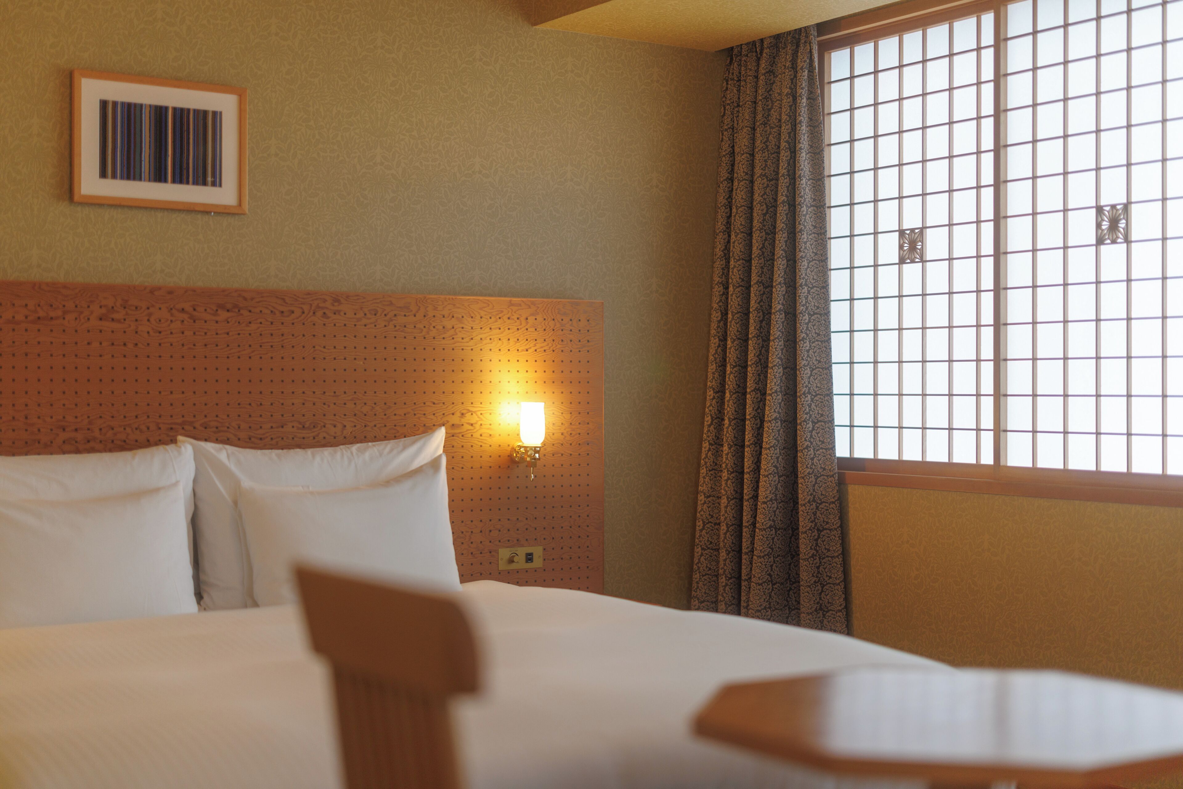 Une chambre d’hôtel avec un lit, une tête de lit, une lampe fixée au mur, une fenêtre avec des rideaux et un tableau encadré accroché au mur.