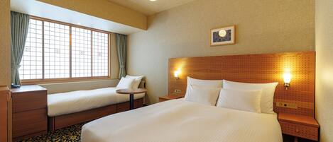 Habitación de hotel con una cama grande, dos camas más pequeñas, cabecera de madera, mesita de noche, lámpara, ventana con cortinas y una alfombra estampada.