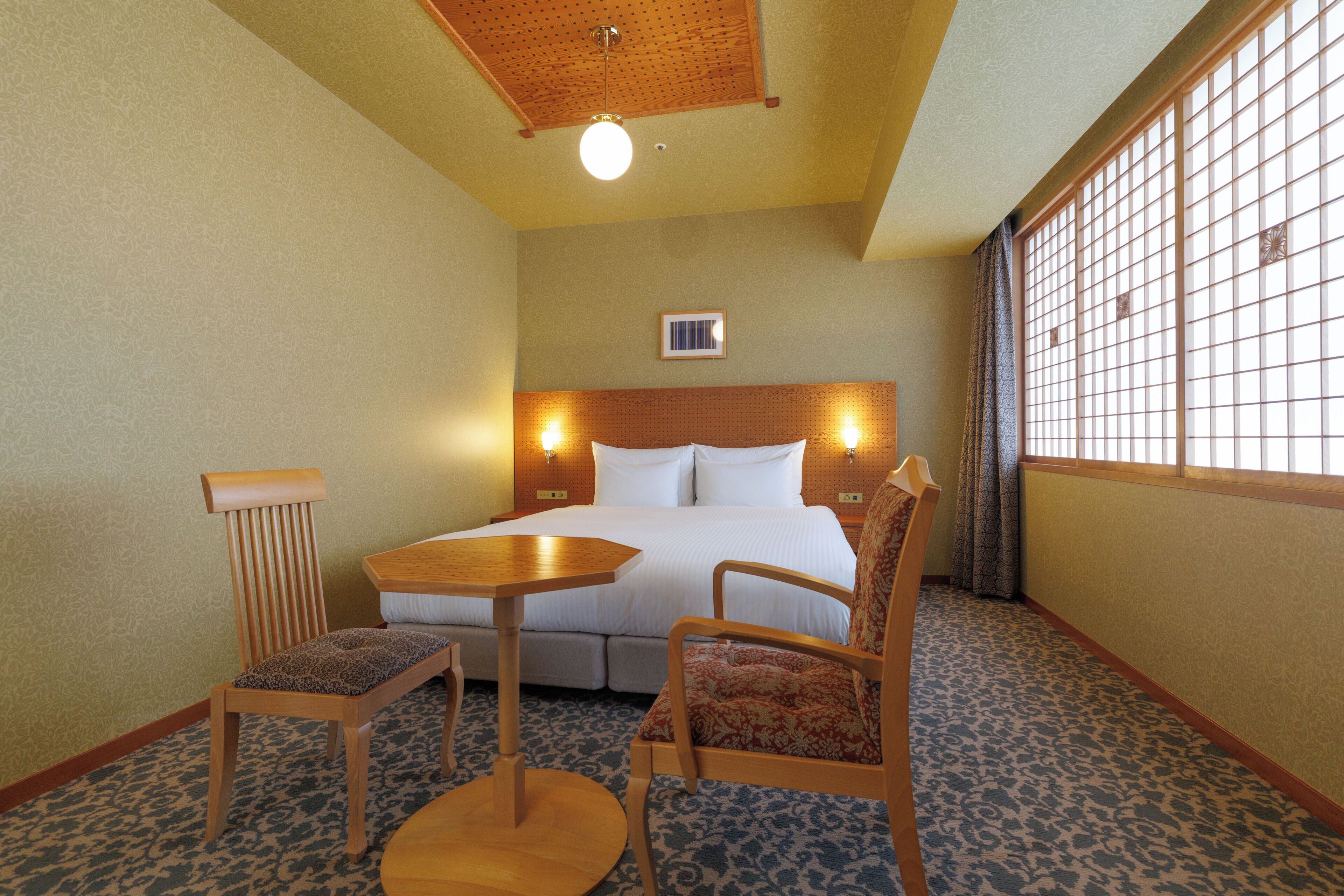 A hotel room with a bed, a wooden table, two chairs, a patterned carpet, and a window with a grid pattern.
