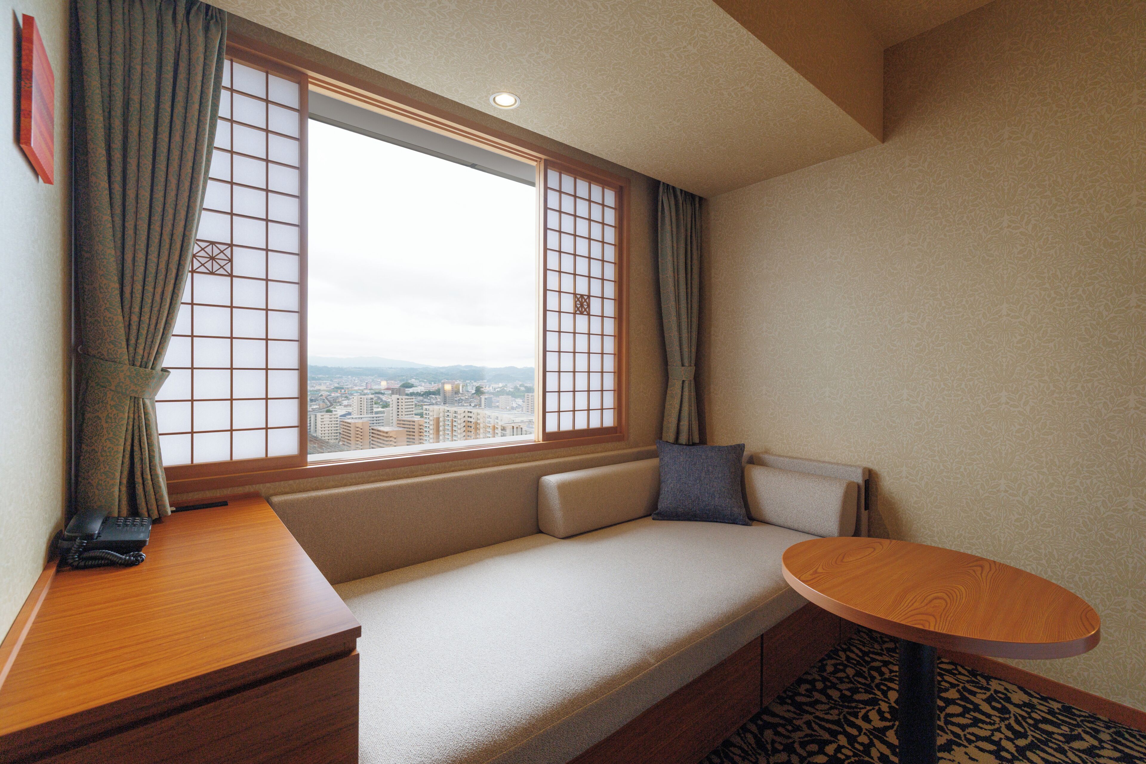A hotel room with a sofa, a small table, a telephone, and a view of the city through a window with a grid pattern.
